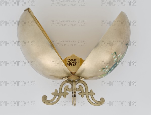 Easter egg, silver, smooth, composed of two opening halves on a hinge, cut vertically; on one part an enameled bouquet of forget-me-nots; gilded interior, with an octagonal basket for embedding a surprise missing. Egg supported on a four-legged stand. Decorated with lilies of the valley, flowers., Date: 1890s, St. Petersburg. Height 11.4 cm; width 6.6 cm; depth 6.6 cm; weight 233.5 g., A metallic, egg-shaped object opened into two curved halves resting on an ornate stand, showing a smooth, slightly tarnished surface in a pale silver tone with subtle darker marks and scratches; the interior rim and decorative base have a warm gold color, and one exterior half features a painted floral motif with green leaves and small white blossoms; the background is a soft, neutral off-white.
