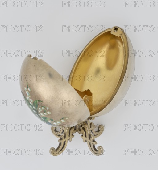 Easter egg, lilies of the valley and flowers motif; silver egg, smooth, composed of two opening halves on a hinge, cut vertically; on one part an enameled bouquet of forget-me-nots; gilded interior with an octagonal basket for embedding a missing surprise. Egg supported on a four-legged stand; 1890s, St. Petersburg; decorative object. Height 11.4 cm, width 6.6 cm, depth 6.6 cm, weight 233.5 g., A decorative egg-shaped object sits open on an ornate stand, showing a shiny gold-colored interior and a muted silver-gray exterior with painted green leaves and small white bell-shaped flowers; the stand and inner rim have warm golden tones, and the background is a pale light gray.