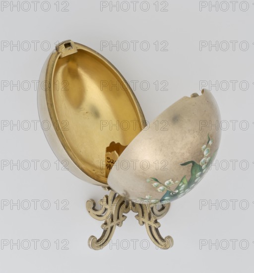 Easter egg, lilies of the valley and flowers motif; silver egg, smooth, composed of two opening halves on a hinge, cut vertically; one half with an enameled bouquet of forget-me-nots; gilded interior with an octagonal basket for embedding a missing surprise. Egg supported on a four-legged stand. 1890s, St. Petersburg. Decorative object. Height 11.4 cm, width 6.6 cm, depth 6.6 cm, weight 233.5 g., An ornate egg-shaped container is shown open on a decorative stand, with the interior displaying a metallic gold color and the exterior a muted silvery-gray with painted green leaves and small pale white flowers; the stand and some interior edges are a warm brass color, and the background is a light neutral gray.