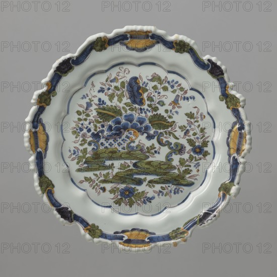 Plate, faience factory Pasquale Antonibon active 1670–1884 maker, reclaimed provenance. Round, shallow plate with irregular wavy rim embossed with 16 semicircular shallow arcades, bordered with two purple lines. Colorfully painted with Far Eastern–style floral and plant decoration in an a ponticello bridge motif; bottom filled with a Rococo composition reminiscent of a Chinese peony bush growing on rocks, created by an openwork structure from behind which emerges a peony bush with delicate twigs, small leaves, and flowers. Colors: purple, light and dark green, yellow, ochre, cobalt, black. Ca. 1750. Nove, Italy. Vessel. Height 2.8 cm; width 24.0 cm., A round decorative plate with a scalloped rim showing a floral scene in the center, featuring a large stylized flower and surrounding foliage, leaves, and smaller blossoms in shades of blue and green with touches of yellow and brown; the central field is bordered by a scalloped inner edge with a blue line, and the rim is ornamented with a ...