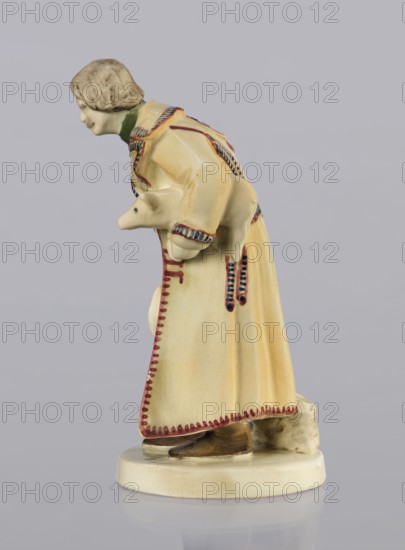 Peasant with Piglet, First Galician Factory of Artistic Faience and Terracotta Pacyków; 1912–1939 manufacturer, Popiel, Antoni 1865–1910 sculptor, figurine of a man peasant, Hutsul russet coat, on a circular base. The peasant, with straight-cut hair and a period mustache, stands slightly bowed, holding a hat in his right hand and a piglet under his left arm. At the back on the base is a woven basket. Warm tones: light ochre russet coat with brown–purple trim; underneath a green vest, dark trousers, and brown shoes. ca. 1910. Pacyków. Terracotta figurine. Height 14.5 cm; width 8.0 cm; depth 5.5 cm., A ceramic figurine of a person wearing a long robe in pale yellow with red and blue painted trim, leaning forward while holding a small animal against their body; the figure has a pale face and short hair shaded in gray, a green collar visible at the neck, and brown shoes peeking from beneath the robe, all standing on a round pale base with a small pale textured object behind the heel.