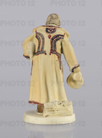 Peasant with Piglet, First Galician Factory of Artistic Faience and Terracotta Pacyków; 1912–1939 manufacturer, Popiel, Antoni 1865–1910 sculptor. Figurine of a man in a peasant Hutsul russet coat, on a circular base. The peasant, with straight-cut hair and a period mustache, stands slightly bowed, holding a hat in his right hand and a piglet under his left arm. At the back, on the base, a woven basket. Warm tones: light ochre russet coat with brown-purple trim; underneath a green vest, dark trousers, and brown shoes. Ca. 1910, Pacyków. Figurine; height 14.5 cm, width 8.0 cm, depth 5.5 cm., A ceramic figurine viewed from behind showing the back of a standing figure wearing a long pale yellow coat with decorative patterns in dark blue and reddish-brown along the shoulders, cuffs, and lower edge, and a pale hat held in one hand; the coat has molded folds and a slightly glossy finish. The figure stands on a rounded off-white base with a small textured rectangular element at the figure’s f...