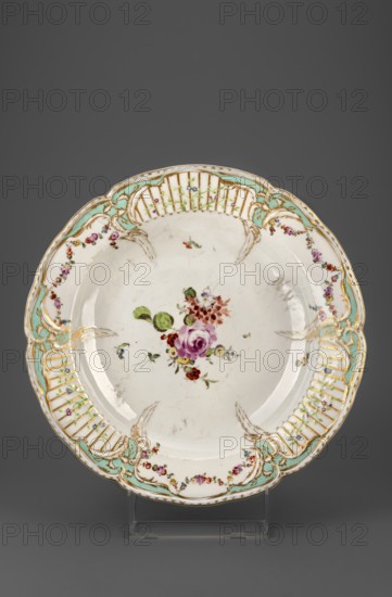 Platter with Reliefzierat decoration and flowers, Königliche Porzellan-Manufaktur KPM, Berlin; maker associated with Frederick II the Great King of Prussia, 1712–1786. Reliefzierat, rocaille ornament; round platter: around the collar, slightly extending into the mirror, a relief of six triangular flamed rocailles with a celadon centre, separating three fields with a trellis of gilded stamens with an interwoven green branch with small flowers Reliefzierat, alternating with three fields with colourful floral decoration and garlands; center of the mirror with a loose floral arrangement, near the rocailles a few small flower sprigs. Half-rolled border with gilding. Ca. 1770–1780, Berlin, Germany. Platter; height 5 cm, depth 34 cm., A round ceramic plate with a scalloped rim displayed on a clear stand against a gray background, the plate having a white base with a central painted bouquet featuring pink, red, and purple flowers with green leaves and small blue accents, the rim decorated with...