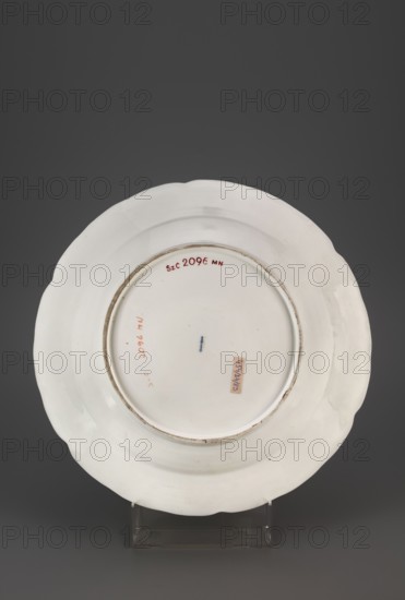Platter with Reliefzierat decoration and flowers, Koenigliche Porzellan Manufaktur KPM; Berlin; 1763–1918. Maker attributed: Frederick II the Great King of Prussia; 1712–1786. Reliefzierat, rocaille ornament, floral decorations. Round platter: around the collar, slightly extending into the mirror, a relief of six triangular flamed rocailles with a celadon centre, separating three fields with a trellis of gilded stamens with an interwoven green branch with small flowers Reliefzierat, alternating with three fields with colourful floral decoration and garlands; in the centre of the mirror a loose floral arrangement, next to the rocailles a few sprays of small flower sprigs. Around the edge, a half-rolled border with gilding. Ca. 1770–1780, Berlin, Germany. Platter; height 5 cm, depth 34 cm., A white ceramic plate is shown from the back, resting on a clear plastic stand against a dark gray background; the plate has a scalloped rim and a slightly raised foot ring, with some small worn or di...