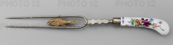 Two-pronged fork with a gilded plastic fish between the tines and a plastic palmette at the base; white metal fork, slightly curved white porcelain handle painted with colorful flower bouquets and insects on both sides. Ca. 1830, Korzec Korzec Porcelain and Faience Manufactory; 1790–1832. Two-pronged cutlery, height 30 cm., A metal two-pronged fork-like utensil with a curved narrow frame and a textured central fish-shaped decoration in gold tone, attached to a metal handle section that transitions into a white curved ceramic grip decorated with painted floral motifs in purple, red, pink, blue, green, and orange, set against a light gray background.