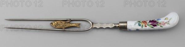 Two-pronged fork made of white metal with a gilded porcelain fish along the length between the tines and a porcelain palmette at the base. Slightly curved porcelain handle, white with colorful painted flower bouquets and insects on both sides. Ca. 1830. Korzec Korzec Porcelain and Faience Manufactory; 1790–1832. Cutlery; height 30 cm. Possible forgery., A metal two-pronged fork with a rounded U-shaped section near the handle holds a small decorative fish figure between the prongs; the metal is silver-colored and the fish is gold-colored with textured scales. The handle is curved and made of white material with painted floral decoration in colors including purple, red, pink, yellow, green, and blue, and there is a metal ferrule connecting the handle to the fork. The background is a neutral light gray.