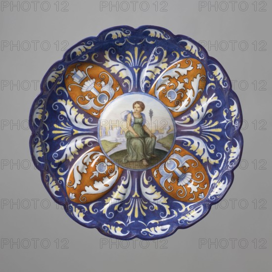 Patera-crespina, Koenigliche Porzellan Manufaktur KPM; Berlin; 1763–1918, imitation Italian majolica, crespina ceramics, Patera - crespisa. On a low hooped foot, modelled-fluted with many leaves, with a slightly raised multi-petaled edge and a raised-convex mirror. The mirror, bordered with a blue string motif, depicts a woman in a green tunic and blue shawl, sitting against a landscape with architecture, with a long arrow stuck in the ground in front of her. Around the collar, on a cobalt background, a motif of four ochre oval palmettes with inscribed decoration with blue plant-ornamental grotesque motifs, alternating with bipartite white-yellowish leafy grotesque motifs on a cobalt background; around the inner edge, a light leafy creeping ornament. Imitation of the form and decoration of 16th-century Italian maiolica a'quartieri from Faenza, 1849–1870, Germany, Berlin. Plateau; height 5.5 cm, width 30 cm., A decorative round plate with a scalloped edge, primarily blue and white with ...