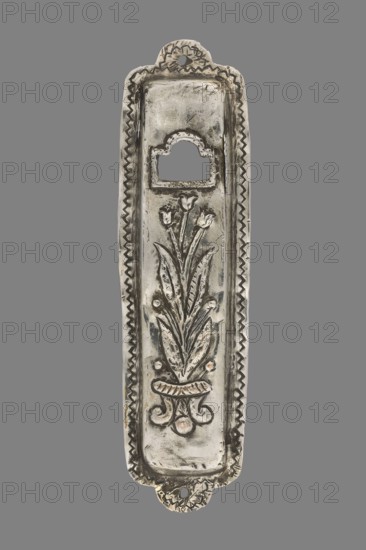 Mezuzah, Judaica; rectangular profiled sheet of low-grade silver edged with a flat ribbon decorated with a zigzag pattern and holes on the longer axis for nailing to a door frame; decorated with a bouquet of tulips in a vase and, above it, a rectangular opening with a semicircular finial; 1st half of the 19th century, Poland, tulip motif, height 10.1 cm, width 2.7 cm., A vertical metallic decorative plate with a long narrow shape displayed against a gray background, showing a raised floral motif in the center with stylized leaves and tulip-like flowers above a small ornamental base, bordered by a zigzag pattern along the inner edges and scalloped decorative tabs at the top and bottom, surface showing areas of darker patina and lighter silver highlights with some warm beige tones in a few spots.