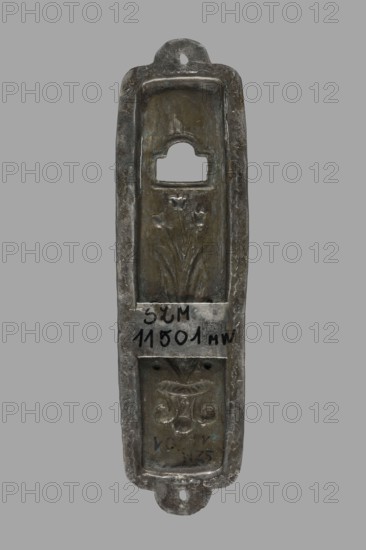 Mezuzah, Judaica; rectangular profiled sheet of low-grade silver edged with a flat ribbon decorated with a zigzag pattern and holes on the longer axis for nailing to a door frame; decorated with a bouquet of tulips in a vase, above it a rectangular opening with a semicircular finial; 1st half of the 19th century, Poland, height 10.1 cm, width 2.7 cm., A narrow vertical metal object with a rounded top and bottom and a central recessed panel, showing a worn metallic surface with areas of dark gray, brownish-green, and lighter silvery gray; the top recess has a rectangular opening with a rounded top edge, the middle recessed area features a floral relief with stems and petals, and the lower recessed area contains a decorative motif resembling a stylized vase and scrolls, all set against a plain gray background.