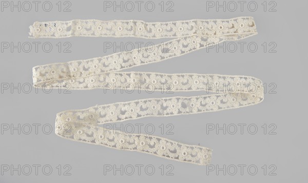 Lace insert, lace, ca. 1920, Poland; clothing; length 133 cm, width 2 cm., A long ornate strip of lace laid out in a zigzag pattern on a flat surface, showing a repeating floral-like openwork motif with circular holes and scalloped edges; the lace appears off-white or cream against a light gray background and shows slight variations in texture and some areas with minor irregularities.