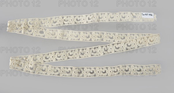 Lace insert, lace, ca. 1920, Poland; clothing; length 133 cm, width 2 cm, A long decorative strip of lace in an off-white or cream color is arranged in a loose zigzag across a pale gray background, showing a repeating floral-like openwork pattern with circular motifs and scalloped edges, with a few areas where the lace overlaps itself and a small rectangular patch of fabric attached near one end.