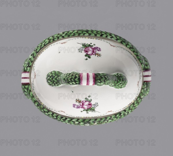 Vase with lid, Prószków Slaski faience factory 1763–1853. Classicist oval vase on a low hooped foot with a tall, domed lid. The rim is framed by a thick plastic laurel garland whose loops hang from the shorter sides to form flat handles; the lid handle is formed from vertically raised scrolls of the garland tied with a three-strand bow. Colorful floral decoration: festoons of small flowers tied with bows and bouquets including a rose, a blooming poppy bud and small flowers. Circa 1770–1783. Origin: Silesia Province of Silesia. Dimensions — vase: height 15.5 cm, width 27 cm, length 36 cm; lid: total height 19 cm, width 21.2 cm, length 30 cm., An oval ceramic lid with a braided handle and rim that appear textured like woven material, predominantly green with darker green shading and touches of black, set against a light gray background; the lid surface is white with a few floral painted decorations in shades of pink, purple, green, and a little orange and blue, and there are bands of pin...