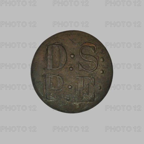 Uniform button, Münchheimer, Ludwika fl. ca 1870–1900 maker, monograms. Round slightly convex uniform button with broken shank; obverse inscribed D:S:/P:E. Reverse rim stamped L. MÜNCHHEIMER / VARSOVIE. Late 19th century, Warsaw, Poland. Clothing accessory, diameter 2.5 cm., A round, flat object with a worn, textured surface in shades of brown, dark green, and muted bronze sits centered on a light gray background; the object's surface shows shallow incised grooves and raised rounded dots, with areas of darker brown patina and small spots of greenish corrosion, and the edges appear slightly irregular and darker than the central area.