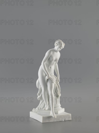 Venus Bathing, after Falconet's sculpture; sculptor: Etienne Maurice Falconet 1716–1791 after Falconet's sculpture; manufacture: Churfuerstliche Porcellain Fabrique, Meissen 1763/74–1814. Figurine of a young, naked woman Venus, long hair parted and arranged in a crown; she stands in contrapposto by a fluted column cut at thigh height on a square plinth, slightly leaning forward, supporting with her hands the heavily draped fabric falling from her left thigh and down the back along the column; she takes a step as if descending from a plinth onto a flat, four-sided base. Marcolini's period 1774–1814. Porcelain sculpture/figurine, 18th/19th century, Meissen, Saxony. Height 25.4 cm; base 10 cm width x 12 cm length., A white sculpted figurine of a nude woman standing on a square base, depicted with a slightly bent posture and looking downward, holding a draped cloth with one hand while the cloth cascades beside and behind her; the figure has curly hair and a smooth, matte surface. The backg...