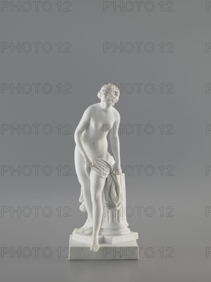 Venus Bathing, after Falconet's sculpture by Étienne-Maurice Falconet 1716–1791; Meissen porcelain figurine, Churfuerstliche Porcellain Fabrique, Meissen Marcolini period, 1774–1814, 18th/19th century, Saxony. Figurine of a young naked woman with long hair parted and arranged in a crown; she stands in contrapposto by a fluted column cut at thigh height on a square plinth, slightly leaning forward, supporting with her hands the fabric falling from her left thigh, heavily draped and falling down the back along the column; she takes a step as if descending from a plinth onto a flat four-sided base. Sculpture porcelain, height 25.4 cm; base 10 × 12 cm., A white marble-like statue of a nude female figure standing on a square base, depicted with a classical pose and curly hair, one arm resting on a fluted column while the other holds a draped cloth; the statue and base are set against a plain gray background with shades of light and darker gray, and the visible colors are white and gray.