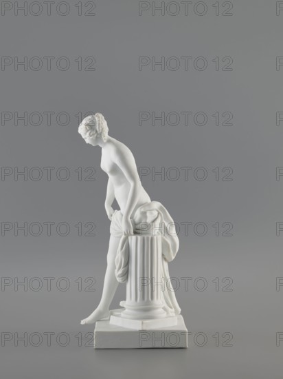 Venus Bathing, after Falconet; sculptor: Etienne Maurice Falconet 1716–1791 after Falconet's sculpture; manufacture: Churfuerstliche Porcellain Fabrique, Meissen 1763/74–1814. Figurine of Venus mythology, Marcolini's period 1774–1814. Porcelain sculpture/figurine, 18th/19th century, Meissen, Saxony. Depicts a young naked woman with long hair parted and arranged in a kind of crown; she stands in contrapposto by a fluted column cut at thigh height on a square plinth, slightly leaning forward, supporting with her hands the fabric falling from her left thigh, heavily draped and falling down the back along the column; she takes a step as if descending from a plinth onto a flat four-sided base. Height 25.4 cm; base width 10 cm; base length 12 cm., A white statue of a nude female figure leaning forward with her hands resting on a fluted cylindrical pedestal draped with cloth, the figure standing on a square base with one foot slightly extended, against a smooth gray background; visible colors...