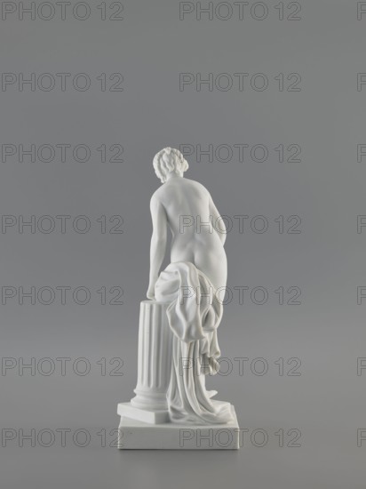 Venus Bathing, after Falconet; sculptor: Etienne Maurice Falconet 1716–1791; manuf.: Churfuerstliche Porcellain Fabrique, Meissen manufacture active 1763/74–1814; figurine of Venus mythology, Marcolini's period 1774–1814; description: young naked woman with long hair parted and arranged like a crown, standing in contrapposto by a fluted column cut at thigh height on a square plinth; slightly leaning forward, supporting with her hands a heavily draped fabric falling from her left thigh and down the back along the column; takes a step as if descending from a plinth onto a flat four-sided base; medium: porcelain sculpture/figurine; date: 18th/19th century; origin: Meissen, Saxony; dimensions: height 25.4 cm, base width 10 cm, base length 12 cm., A white marble-like sculpture of a nude figure seen from the back, standing with one hand resting on a fluted column draped with a flowing cloth, on a square base; the figure has short, curly hair and smooth contours. The background is a uniform g...
