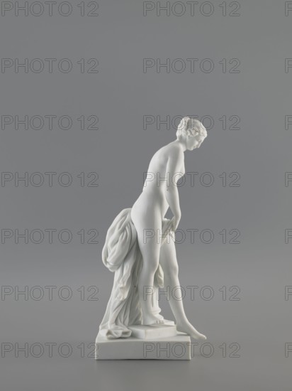 Venus Bathing, after Falconet; sculptor: Étienne Maurice Falconet 1716–1791; manuf.: Churfuerstliche Porcellain Fabrique, Meissen 1763/74–1814, Marcolini's period 1774–1814. Figurine of Venus mythology: a young naked woman with long hair parted and arranged in a crown; standing in contrapposto by a fluted column cut at thigh height on a square plinth, slightly leaning forward, supporting with her hands a heavily draped fabric falling from her left thigh and down the back along the column; she takes a step as if descending from a plinth onto a flat, four-sided base. Porcelain sculpture, 18th/19th century, Meissen, Saxony. Height 25.4 cm; base width 10 cm; base length 12 cm., A white statue of a nude female figure standing on a white rectangular base, shown in profile and leaning slightly forward with a relaxed posture, draped with a flowing cloth gathered behind her and hanging toward the base, set against a plain gray background; visible colors include white and shades of gray.