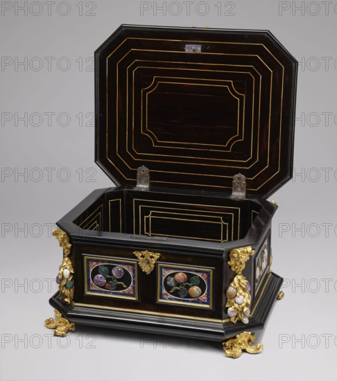 Jewel box, pietra dura technique; made of blackened wood, partly ebony; cuboid with chamfered corners supported on four gilded bronze volute legs decorated with a mascaron at the base. Edges of base and lid with profiled ebony strips. Decorated on side walls with six mosaic reserves and one plaque on the lid: front and back panels show two fruits on a branch; side panels show three fruits on a branch and an insect at the bottom. Corners feature cast gilded bronze garlands topped with medusa masks and plastic fruits made of semi-precious stones 7 pieces each. Hinged, profiled lid with a large plate showing three fruits on branches tied with a sash and an insect above; side walls decorated with gilded bronze garlands with plastic fruits; corners with gilded bronze shells; front with preserved keyhole. Dated to the early 18th century; attributed to Florence, Italy ; chest; height 34.5 cm, width 41.5 cm, depth 50.5 cm., A dark wooden octagonal chest with its lid open, showing concentric re...
