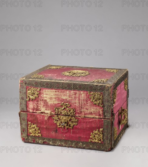 Casket case. Rectangular wooden box covered with red velvet, with gold thread trimmings on the edges. Crosspieces have bronze openwork overlays. Side walls have gilded bronze handles; top has a decorative overlay in the middle with a rank crown. Front wall has a decorative overlay serving as a lock plate; keyhole preserved. Interior lined with light green silk. Early 19th century; 18th century, Italy, Florence. Chest. Height 39.5 cm; width 56.7 cm; depth 47.5 cm., A worn rectangular box covered in faded red fabric with decorative dark trim and a group of ornate metal appliques in a gold color placed at corners and centers; the fabric shows areas of wear with lighter, scraped sections and frayed edges, and a few strands of fiber are visible along a seam; the trim has a textured, patterned appearance in a dark grayish tone, and there is a metal handle in a gold color on one side; the background is a smooth light gray surface and backdrop.