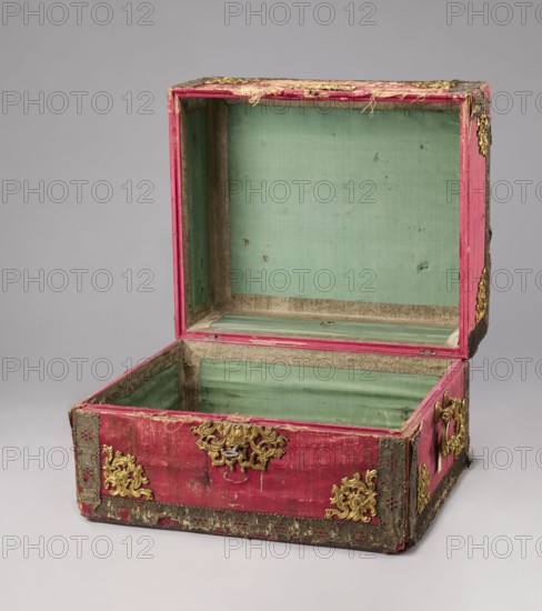 Casket case. The case is a rectangular wooden box covered with red velvet, with gold thread trimmings on the edges. Crosspieces have bronze openwork overlays. Side walls have gilded bronze handles; the top has a decorative central overlay with a rank crown. The front has a decorative overlay serving as a lock plate; the keyhole is preserved. Interior lined with light green silk. Early 19th century; 18th century, Italy Florence?. Chest. Height 39.5 cm, width 56.7 cm, depth 47.5 cm., An open rectangular box with a hinged lid, the exterior covered in worn pink fabric with areas of fraying and darker aged spots, embellished with ornate gold-colored metal corner and decorative fittings; the interior lined in a pale green fabric with visible wear and small blemishes, and inner edges trimmed in a muted beige material; the box sits on a neutral light gray surface against a matching gray background, showing the lid propped open and the empty interior visible.