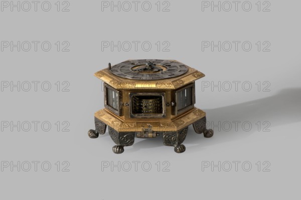 Tiled clock; maker: Betzmeier, Erasmus fl. ca. 1750, clockmaker; mid-18th century, Warsaw; materials/ornament: acanthus ornament, lion's paws; function: timepiece; dimensions: height 8 cm, width 12.8 cm, An ornate hexagonal box-like object with a metallic appearance sits on a flat surface against a plain background; it has a gold-colored top rim and base with engraved decorative patterns, dark gray circular elements and mechanisms on the top surface, glass panels set into the sides revealing inner components, and dark silver or black sculpted feet with rounded ends supporting the piece, with visible wear and patina on several surfaces and a soft shadow extending to one side; colors visible include gold, dark gray, black, silver, and transparent glass.