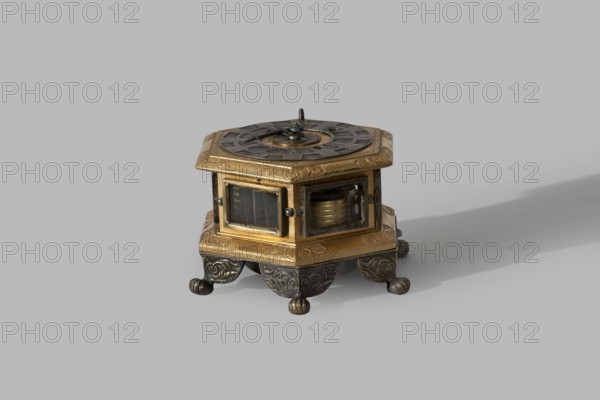 Tiled clock, Betzmeier, Erasmus fl. ca. 1750 clockmaker, acanthus ornament, lion's paws, mid-18th century, Warsaw, timepiece, height 8 cm, width 12.8 cm, A small hexagonal mechanical box sits on a smooth neutral background, featuring a mix of metallic gold and darker aged metal tones. The top is decorated with a circular patterned plate in a darker metal with a small central pin and radial markings, while the surrounding top edge is gold with engraved ornamental designs. The sides have glass panels revealing internal gears and a cylindrical mechanism in a brass tone, framed by gold borders and darker metal fastenings. The base is gold with engraved decoration and rests on ornate dark metal feet with scrollwork and rounded end caps. Shadows fall softly to one side across the pale background, and visible colors include gold, brass, dark grey, black, and the pale neutral of the backdrop.
