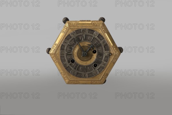 Tiled clock, Betzmeier, Erasmus fl. ca. 1750 clockmaker, acanthus ornament, lion's paws, mid-18th century, Warsaw, timepiece, height 8 cm, width 12.8 cm, A hexagonal metallic object with a central circular face is visible, showing a mix of gold and dark gray tones; the outer hexagon frame is gold with engraved decorative patterns, and the inner circular rings are dark gray with textured segments surrounding a small gold circular center that also has engraved designs; a group of dark bluish-gray hands and small circular openings appear on the central face, and small rounded dark knobs protrude from the hexagon edges; the background is a smooth gradient of light gray to medium gray.