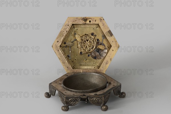 Tiled clock, Betzmeier, Erasmus fl. ca. 1750 clockmaker, acanthus ornament, lion's paws, mid-18th century, Warsaw, timepiece, height 8 cm, width 12.8 cm, An ornate hexagonal metal object with a hinged top opened to reveal an intricate inner mechanism; the top interior is a brass-colored plate with decorative filigree elements, small dark screws, and engraved ornamental parts, while the outer edges of the lid show areas of darker tarnish and worn spots in brownish tones. The base is a darker silver-gray metal bowl set into a square platform with rounded corners, supported by four short, ball-shaped feet with carved swirl patterns and a patina of dark gray and brown. The visible colors include brass yellow, silver-gray, dark gray, brown, and hints of black on small fasteners and shadowed recesses.
