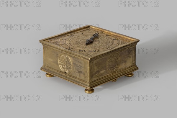 Tiled Clock, acanthus ornament, plant tendril, ca. 1580, Germany, clock, height 6.3 cm, width 13.4 cm, depth 13.4 cm, A square ornate metal box with a golden-brass finish sits on a flat surface, supported by small rounded feet; the top features a circular engraved design and a decorative dark metal hand or pointer lying across the center, and the sides display circular floral medallions and rectangular panels with engraved detailing. The visible colors are gold, brass, dark brown, and grey.