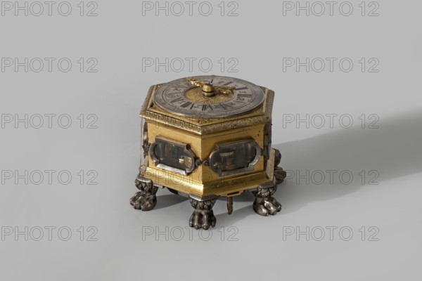 Tiled clock, Zoll, Beniamin fl. 1715–1760 clockmaker, lion's paws, plant tendril, winged putti iconography, ca. mid-18th century, Gdansk, Poland; Royal Prussia; clock; height 9.1 cm, width 13.1 cm, depth 9.6 cm, A small hexagonal mechanical object sits on a smooth surface, featuring a metallic top with a circular dial and ornate central knob, a body made of a gold-colored metal with decorative edges and inset framed windows on its sides, and a base supported by a group of sculpted lion-paw feet in a darker silver-gray metal; visible colors include gold, silver-gray, and muted gray of the background, with subtle darker shadows cast to one side.