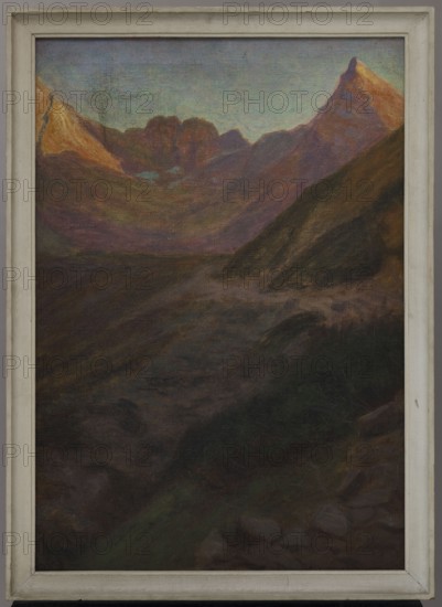 Sunset in the Mountains, Ostrowski, Wladyslaw 1878–1942, painter; 1913, oil painting, 80 × 50 cm., A framed landscape painting showing a mountainous scene with a prominent peak on the right and a range of jagged peaks toward the center and left; the sky above the mountains appears in pale blue and teal tones, while the upper slopes of the peaks display warm oranges and muted purples; the middle ground contains rolling dark ridges in deep purples, browns, and greens, with a sloping dark hillside descending from the right; the foreground is shadowed with dark browns, deep greens, and near-black areas suggesting rocky terrain; the painting is surrounded by a light-colored wooden frame in off-white and pale gray.
