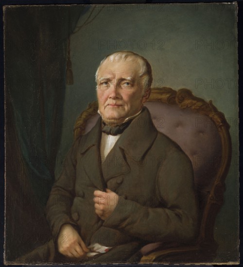 Portrait of Stanislaw Kurpiewski; painter: Rafal Hadziewicz 1803–1886. Ca. 1860. Oil painting, 58 x 53 cm. Half-length, three-quarter, armchair portrait of a seated grey-haired, balding older man; interior setting with curtains. Inscription/subject: Stanislaw Kurpiewski. Genres: male portrait, old-age portrait., An older man with pale skin and white hair sits in a carved wooden chair upholstered in a muted purple fabric, wearing a dark brown coat and a black neckcloth over a white shirt; his expression is neutral and he looks slightly upward and to his left. His left hand rests on his lap holding a white handkerchief with a small colored spot, and his right hand is positioned near his chest clutching his coat. Behind him is a backdrop of dark green drapery at the left and a soft teal wall at the right, with warm brown and gold tones in the chair frame and subtle highlights on his face.