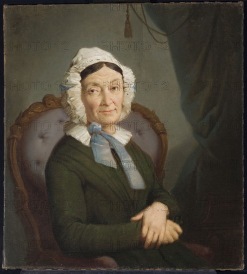 Portrait of Anna Kurpiewska, née Olszakiewicz; painted by Rafal Hadziewicz 1803–1886. Oil painting, ca. 1860; height 58 cm, width 53 cm. Half-length, en face portrait of an elderly seated woman in an armchair, wearing a bonnet and curtains visible in the interior. Portrait, female portrait., An older woman sits in an upholstered wooden chair, wearing a white ruffled cap with light blue ribbons and a dark green dress; her hands are folded in her lap and her face shows a neutral expression with visible wrinkles and pale skin tones. The chair has a carved wooden frame and a tufted back in muted purple. The background includes a draped dark curtain with teal and dark green shadows and a lighter gray wall area behind her. Colors visible include white, light blue, dark green, muted purple, brown, gray, teal, and flesh tones.