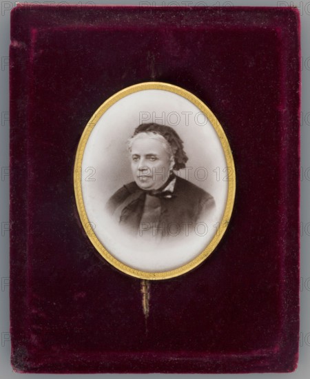 Portrait of Mlle Rousset at about age 70 died 1893, secretary and librarian of Izabella; photographer: Braun, Adolphe 1812–1877 author; from 1889 Braun, Clément et Cie, and 1910 – Braun e; inscription/note: Josephine Rousset, niece of the Czartoryski doctor younger than Izabella, a student of the Hotel Lambert, a companion of the Czartoryskis' youth, over time gathered in her hands the administrative threads of Izabella's activities, and partly of Wladyslaw Czartoryski's.; dated 1870; place of creation: France; miniature; height 6.8 cm, width 5.4 cm; en face portrait of a woman wearing headgear., An oval miniature portrait of an older person with light skin and short hair, wearing a dark garment and a dark head covering, set within a thin gold frame; the portrait sits against a textured deep burgundy velvet background with visible wear and small fiber details, and a narrow strip of gold beneath the frame.