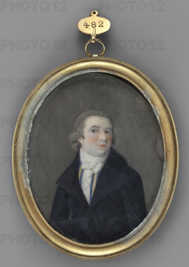 Portrait of a Man in a Black Frock Coat, Legrand, Franz 1807–1833 painter. Subject: men's portrait, 3/4 right, to the waist; young man with ash-grey hair and grey eyes. Costume: white cravat, vest with blue and white stripes and white checkered front, black frock coat. Medium/type: miniature. Binding: oval rim made of gilded brass, cylindrical, with a braided motif in the center. Date/place: 1850, Germany. Size: height 8.4 cm, width 6.7 cm., An oval portrait set in a gold-colored metal frame with a small loop and hanging tag at the top shows a man with light skin and shoulder-length brown hair wearing a dark coat and a white cravat; his face has pale skin with pinkish cheeks and he looks slightly to one side against a muted gray background, with visible wear and discoloration along the inner edge of the frame and a few blue and yellow accents on the man's clothing.