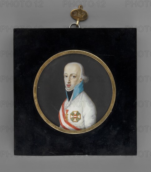 Charles Louis of Habsburg 1771–1847, son of Leopold II and Maria Louise of Bourbon. Johannes Moltzheim 1771–1825 painter. Male portrait, bust left. Light blond/almost white hair, blue eyes. Wearing white uniform with blue collar, white shirt hem and black scarf; white and red ribbon; the Order of the Golden Fleece on the ribbon and the cross of the Order of Maria Theresa on the left breast. Background dark gray. Narrow medallion rim of gilded bronze. 1805, Austria. Miniature, height 6.8 cm., A small framed circular portrait set in a rectangular black panel shows a three-quarter view of a pale-skinned man with light-colored hair styled back from his forehead. He wears a high blue collar over a dark inner garment and a white outer coat with a diagonal red-and-white sash across his chest. A decorative cross-shaped medal with green and gold elements is pinned on his coat. The circular portrait is bordered by a gold rim, and the black panel has a small gold hang tag attached at the top. Col...