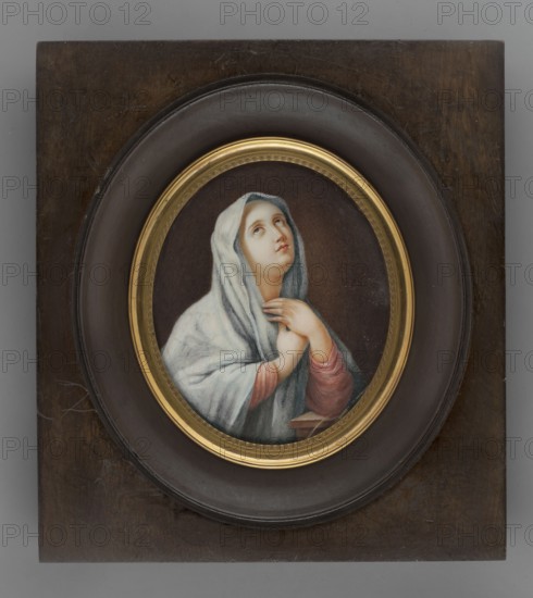 Mary Magdalene — copy from Guido Reni; painter: Thil, Wilhelmine Louise du 1782–1819. Portrait of a woman in dress à l'antique with headscarf; miniature, 1820. Height 9.1 cm, width 7.1 cm., An oval framed oil painting set into a dark wooden rectangular panel shows a woman wearing a pale blue-gray head covering and a light garment, her face turned upward with a contemplative expression, visible skin tones of peach and pink on her face and hands, with her hands clasped near her chest and sleeves of muted pink beneath the outer drapery; the oval frame has a thin gold inner rim and sits within a broader dark brown circular mount against the wood panel; visible colors include blue-gray, pale pink, gold, dark brown, peach, and black.