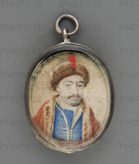 Portrait of an unknown man in oriental costume. In almost straight half-length, head slightly to the right. He wears a sable cap with a red tuft on the front tuft painted later, per conservator analysis, a sapphire zupan, a white belt with red stripes, and on the shoulders a red delia with a fur collar and lapels. Background light. Frame: border of silver braided wire on a silver rim with traces of gilding, framing two thick, beveled outer glasses. On the reverse, on cardboard, a 20th-century ink inscription crossed out: Bohdan Chmielnicki. It is very doubtful that the identification as Chmielnicki is truthful—the physiognomy does not resemble the figure from the well-known 1651 engraving by Willem Hondius and the costume does not resemble Cossack clothing; pre-war research suggested the image may depict an unknown Turk. Miniature, after 1800?, height 3.1 cm, width 2.6 cm., An oval pendant with a metal loop at the top contains a small painted portrait of a person wearing a brown fur ha...