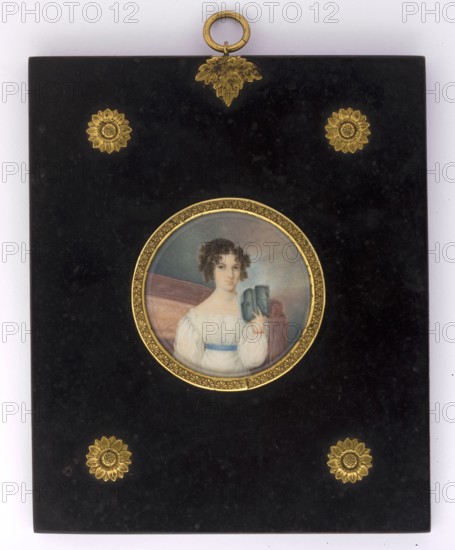 Portrait of a woman reading a book, unknown author amateur; books; woman; full-face portrait; interior; female portrait; hairstyle; Polish miniature; amateur; 1825; miniature, A rectangular black plaque with a glossy finish features a central round miniature portrait set in a decorative gold frame; the portrait shows a person with curly dark brown hair wearing a white dress with a blue sash and holding a small blue-gray object, seated against a muted background of soft gray and brown tones. The plaque is embellished with several ornate gold floral or sunburst motifs near the corners and a gold loop and leaf-shaped ornament at the top. The visible colors include black, gold, white, blue, brown, gray, and subtle flesh tones.