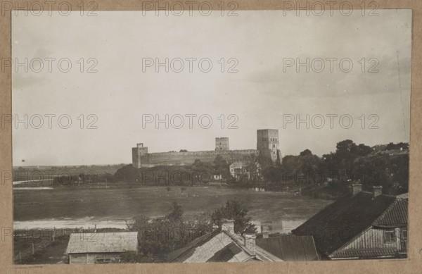 Lutsk. Lubart's Castle – general view from the Styr River, unknown creator/photographer, architecture, castles; Polish architecture; Lutsk Ukraine; photographic print glued to brown cardboard; ca. 1918 negative, ca. 1918–ca. 1922 print; nature photography/photography; photographic print: height 10.2 cm, width 15.9 cm; cardboard: height 35.5 cm, width 25.5 cm., The image shows a grayscale scene with a large stone fortress or castle with a few tall rectangular towers situated across a body of water, surrounded by trees and low buildings; in the foreground there are rooftops of structures with shingles and chimneys, and a few trees and fences near the water’s edge. The sky is light gray with some darker cloud patches, and the overall colors visible are shades of gray, black, and muted brown tones from the mounted border.