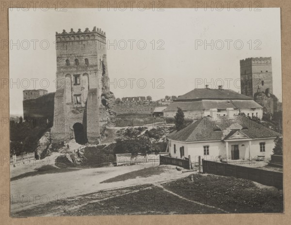 Lutsk. The Castle – view towards the Styrowa Swidrygiello Tower, /photographer, architecture, Polish architecture, Lutsk Ukraine, castles, ca. 1918 negative, ca. 1918–ca. 1922 print, Lutsk, photography, nature photography, photographic print: height 11.2 cm, width 14.6 cm; cardboard: height 35.1 cm, width 25.2 cm., The image shows an outdoor scene with a large stone tower ruin and sections of a fortified wall alongside a group of low buildings with pitched roofs; the tower and wall appear in shades of gray and brown, and the buildings are in lighter gray and off-white tones with darker gray roofing. In the foreground there is an unpaved area and a grassy patch in muted greenish-brown, bordered by a dark wooden fence and a few scattered shrubs and small trees in dark green. A path and a simple wooden gate lead toward the base of the tower, and there are several trees and low vegetation between the structures. The sky is pale and almost white, creating a high-contrast backdrop to the arc...
