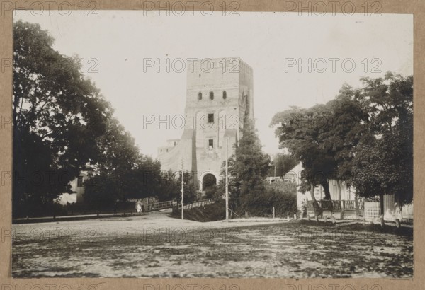 Lutsk. Castle – view towards the Lubart Tower, unknown creator/photographer; architecture; Polish architecture; castles architect; Lutsk Ukraine; photograph glued over entire surface to a museum backing together with another photo; negative ca. 1918, print ca. 1918–ca. 1922; photography, nature photography; photographic print: height 11.0 cm, width 16.5 cm; cardboard: height 35.1 cm, width 25.2 cm., A sepia-toned photograph showing a large stone tower centered in the scene, flanked by walls and trees on either side. In the foreground there is a wide open dirt or paved area with a textured surface. A small bridge or walkway leads toward the arched entrance at the base of the tower. On both sides of the tower are dense groups of trees and foliage in dark green tones, with lighter green bushes nearer the entrance. A few small buildings with pale walls and darker roofs are partially visible among the trees on the right and left. Several vertical poles or posts stand near the bridge. The ov...