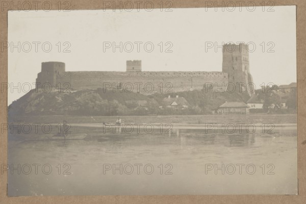 Lutsk. Lubart's Castle – general view from the Styr river, unknown author/photographer; architecture; castles; Polish architecture; Lutsk Ukraine; Styr river; rivers. Photographic print glued to brown cardboard. Negative ca. 1918; print ca. 1918–1922. Place of creation: Lutsk. Photography from nature. Photographic print: height 12.1 cm, width 20.6 cm; cardboard: height 35.3 cm, width 25.4 cm., The image shows a wide, muted scene with a large stone fortress or castle stretching across a raised hillside beside a body of water; the fortress has a few tall towers and long curtain walls in shades of gray and brown, with the hillside beneath it covered in darker greenish-gray vegetation. In front of the hill, there is a riverside area with several small buildings and houses in pale gray and off-white tones, and a few trees with darker green-gray foliage. On the water there are a couple of small boats, one with a person seated and another with a person standing, rendered in dark gray silhouet...