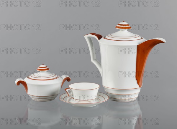 Jug with a lid from the Baltyk set, designed by Bogumil Marcinek 1904–1940, manufactured by Fabryka Porcelany i Wyrobów Ceramicznych Cmielów SA ca. 1930. White porcelain, glazed, decorated in form and partly on the surface with orange overglaze paint. The jug is slightly widened toward the top and flared like a goblet, enriched in the lower part with three concave, arched recesses narrowing the vessel, highlighted with orange lines painted overglaze. The decorative shape of the lower part is repeated in the lid’s handle, where the folds are highlighted with orange circles. The jug’s handle echoes the concave folds of the body, creating a cup-like form that tilts outward while the upper, enclosing part has three semicircular protrusions; the outer part of the handle is painted orange. Vessel; height 19.0 cm, width 22.1 cm., A group of white ceramic tableware pieces including a teapot with a tall body and a curved spout, a lidded sugar bowl with two curled handles, and a teacup resting o...