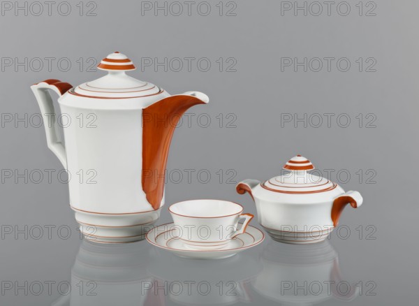 Cup from the Baltyk set, designed by Bogumil Marcinek 1904–1940, manufactured by Fabryka Porcelany i Wyrobów Ceramicznych Cmielów SA c. 1920–1951. White fine porcelain cup, glazed; bowl widens and flares outward at the top, narrows at the bottom with three concave embossments. Rim and lower bends emphasized by thin orange lines. Handle echoes the concave bends, forming a cup-like outward bend; upper part delicately fluted, matching the jug and sugar bowl from the set. Outer part of the handle painted orange. Circa 1930., A white porcelain tea set sits against a gray background, consisting of a tall teapot with a domed lid and a curved handle, a covered sugar bowl with two handles and a domed lid, and a teacup on a saucer; each piece features orange accents including a broad orange panel on the teapot spout and matching orange on the handles and lid tops, and thin orange concentric rings decorating the lids, rims, saucer, and bases, with reflections visible on the glossy surface and the...