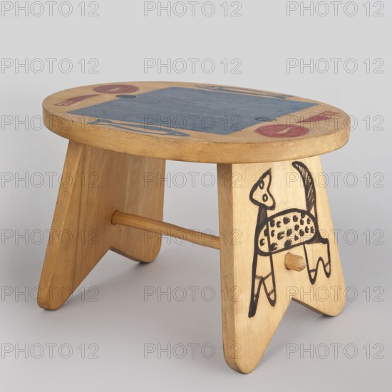 Table from a set of doll furniture. Wooden table with an oval top resting on stringer supports, connected with a lower link. Painted decoration: schematic silhouettes of animals on supports and tableware arrangement on the tabletop. Toy. Maker: Kenar, Antoni 1906–1959, sculptor; co-creators: State Secondary School of Fine Arts in Zakopane Zakopane; 1876–, Office of Production Aesthetics Supervision Warsaw; 1947–1949. Date: 1947–1948. Place of origin: Zakopane. Dimensions: height 12 cm, width 18 cm, depth 15 cm., A small wooden stool with a rounded top and two triangular legs joined by a dowel, the wood showing a light tan color with visible grain; the top surface has a central rectangular area painted blue and a few circular and elongated painted shapes in red and blue near the edges; one leg features a simple black line drawing of an animal with a spotted body and a raised tail; the background is a plain light gray.