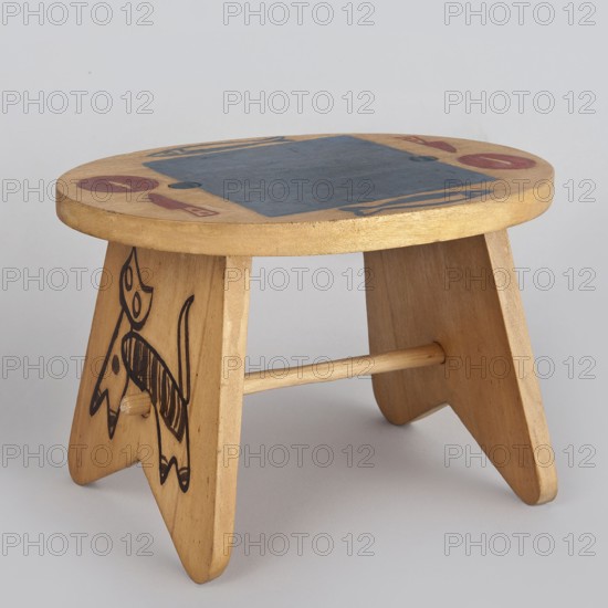 Table from a set of doll furniture, State Secondary School of Fine Arts in Zakopane Zakopane; 1876-, co-creator, Office of Production Aesthetics Supervision Warsaw; 1947-1949 co-creator, Kenar, Antoni 1906-1959 sculptor/author, Wooden table with an oval top resting on stringer supports, connected with a lower link. Painted decoration—schematic silhouettes of animals on supports and tableware arrangement on the tabletop. 1947–1948, Zakopane; toy. Dimensions: height 12 cm, width 18 cm, depth 15 cm., A small wooden stool with a round top and three angled legs joined by a horizontal dowel, showing light tan and beige wood tones with areas of darker brown grain; the top surface has a large dark blue painted rectangle and small circular and crescent-shaped motifs in red and blue, and one leg features a black hand-drawn animal-like figure with bold outlines and striped detail.