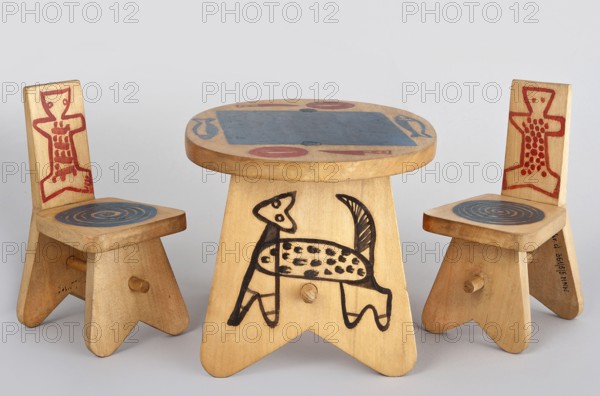 A stool-type chair from a set of doll furniture with painted decoration on the backrest schematic figure of a teddy bear and on the seat circular ornament. Made 1947–1948 in Zakopane. Materials: wood; height 14.0 cm, width 7.5 cm, depth 8.5 cm. Designed by Antoni Kenar 1906–1959. Co-creators: Office of Production Aesthetics Supervision Warsaw; 1947–1949 and State Secondary School of Fine Arts in Zakopane. Function: toy., A small wooden table and two matching chairs arranged on a neutral surface, all made from light-colored wood with simple carved shapes and rounded edges; the table has a round top with a large painted rectangular patch in blue and a few circular and elongated painted motifs in red and blue around its edge, and its pedestal base shows a stylized animal-like figure painted in black on the front; each chair has a tall back with a red, geometric, human-like figure painted on it and a square seat topped with a concentric circular motif in blue, and both chairs have short, s...