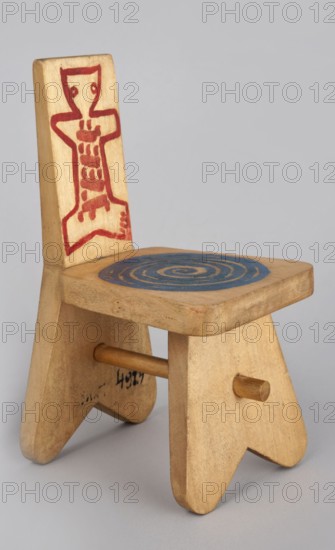 A stool-type chair from a set of doll furniture with painted decoration on the backrest schematic figure of a teddy bear and on the seat circular ornament. Made of wood. Maker: Kenar, Antoni 1906–1959, sculptor; co-creator: Office of Production Aesthetics Supervision Warsaw; 1947–1949; State Secondary School of Fine Arts in Zakopane Zakopane; 1876– . Date: 1947–1948. Place of origin: Zakopane. Object type: toy. Dimensions: height 14.0 cm, width 7.5 cm, depth 8.5 cm., A small wooden chair with a light natural wood finish is shown against a pale gray background; the backrest features a red painted abstract figure design, the seat has a spiral pattern painted in blue, and the legs are simple triangular shapes joined by a round wooden dowel; visible colors include light beige wood, red, blue, and gray.